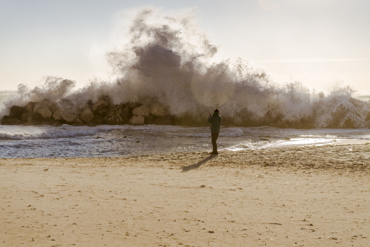 Un homme fait face téléphone en main à une vague qui se brise sur les rochers, pour prendre en photo cette gerbe d'eau sur la plage du Prophète à Marseille
