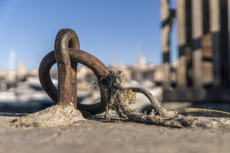 Un anneau d'amarrage rouilé avec une corde effilochée encore nouée, photographié au ras du sol sur le Vieux-Port de Marseille. 