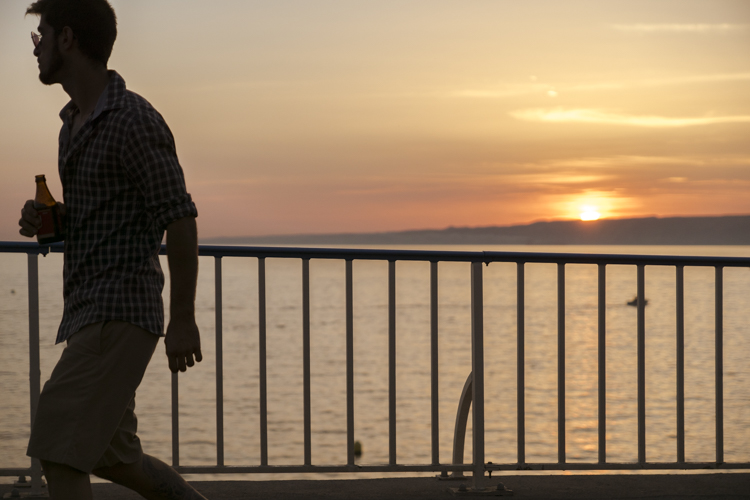 Un homme marche le long de la Corniche Président John Fitzgerald Kennedy à Marseille au soleil couchant et une bière à la main.