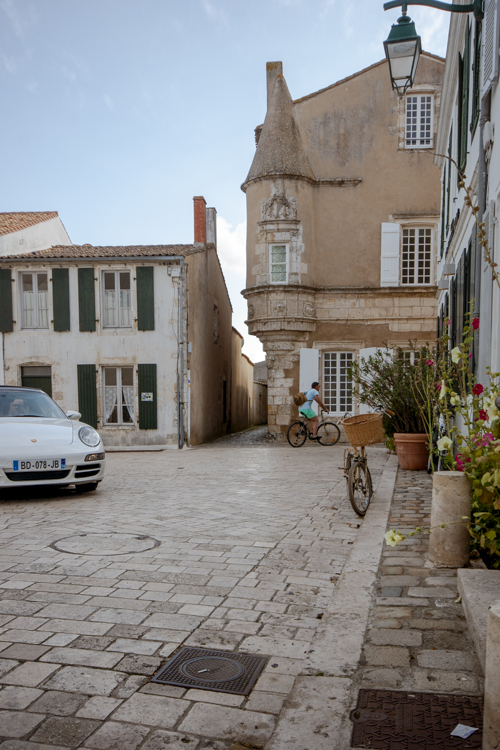 Une rue d'Ars-en-Ré en fin d'après-midi où se croise véhicule de luxe et vélo devant une ancienne bâtisse.